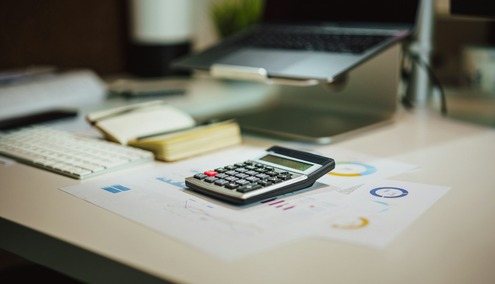 Une calculatrice repose sur des documents financiers accompagnés de graphiques, mettant en avant la facturation électronique, à côté d'un cahier ouvert, d'un clavier et d'un ordinateur portable posé sur un support, dans un bureau au style moderne.
