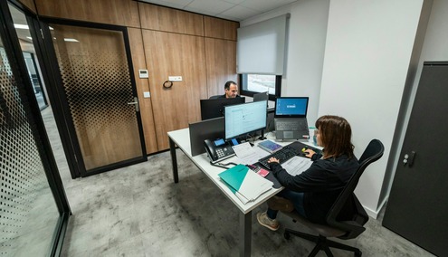 Deux personnes travaillant à leur bureau dans un espace de travail moderne : l'une utilise un ordinateur près de la fenêtre, tandis que l'autre manipule des documents et un ordinateur portable. Cet espace de travail, idéal pour un prestataire de services gérés (MSP) ou une équipe informatique interne, est doté de cloisons en bois, d'une porte vitrée et d'un classeur.