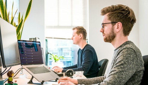 Deux personnes travaillent devant des ordinateurs dans un bureau lumineux. L'une d'elles utilise un ordinateur portable posé sur un support au premier plan, tandis que l'autre, membre de l'équipe MSP PME Belgique, travaille sur un ordinateur de bureau près d'une fenêtre et de plantes vertes.