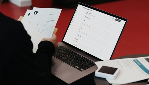 Une personne est assise à une table devant un ordinateur portable ouvert sur un formulaire, tenant des documents imprimés — peut-être en train de se renseigner sur l'automatisation comptable. Un smartphone et d'autres papiers se trouvent sur la table, le tout sur fond rouge.