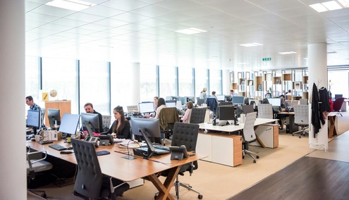 Un bureau en open space où plusieurs personnes travaillent à leur poste dans un environnement informatique, avec de grandes baies vitrées laissant entrer la lumière naturelle et du mobilier de bureau moderne réparti dans tout l'espace.